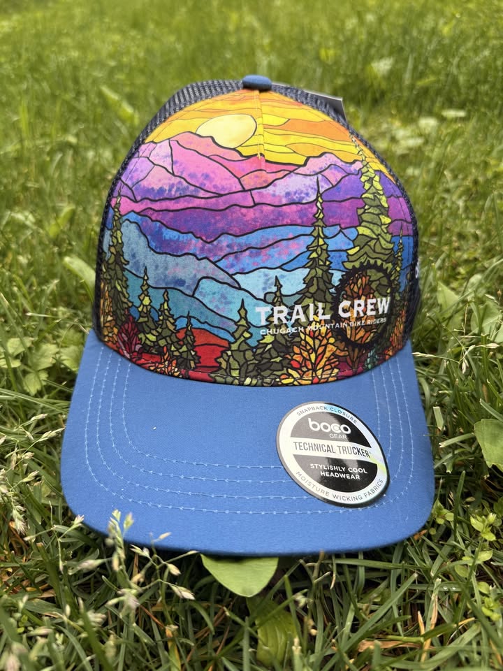 CMBR Trail Crew hat - stained-glass Chugach mountain design on a blue trucker cap, lying in grass