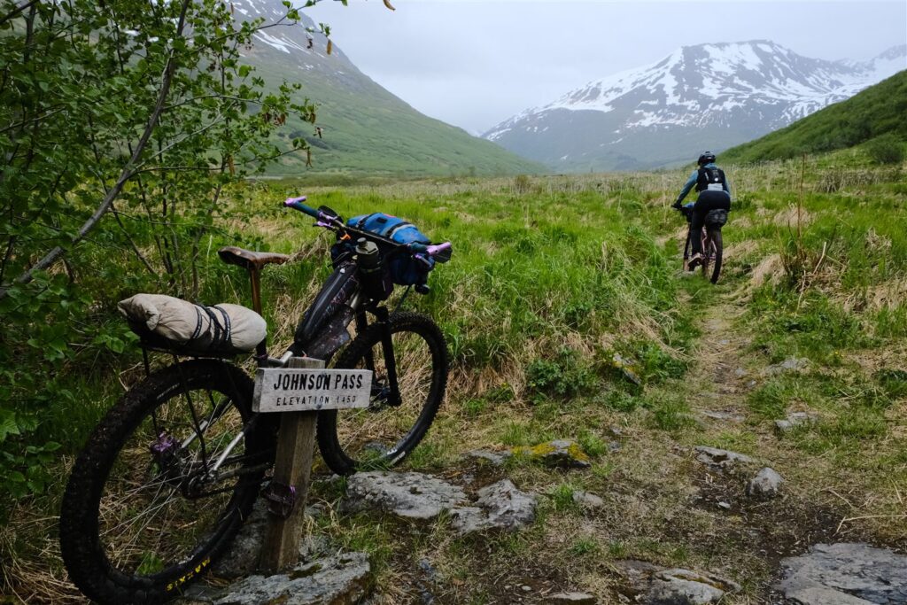 A loaded bikepacking bike leans on the Johnson Pass trail sign, elevation 1450, as a rider pedals up a grassy valley toward snowy peaks.